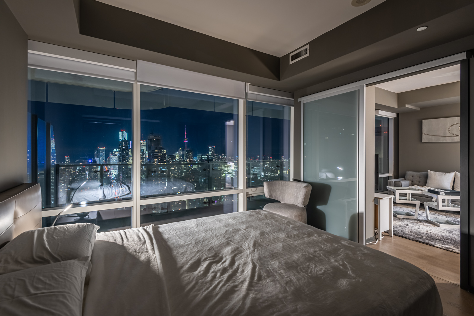 1 Bloor St E Unit 4305 master bedroom at night with 1 lamp; view of Toronto skyline, bright buildings and CN Tower.