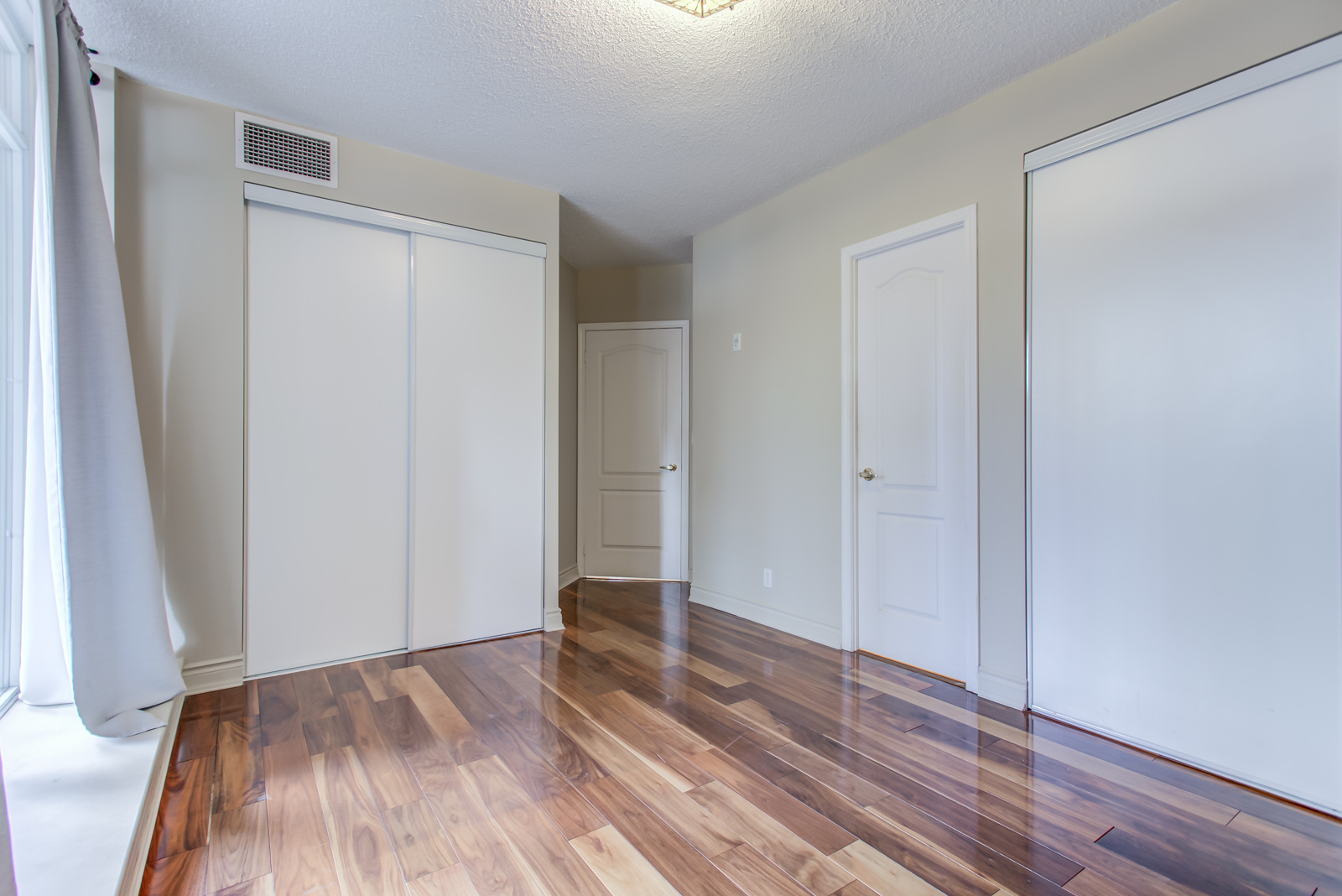 Empty master bedroom with dark and shiny hardwood floors, gray walls, and multiple doors.