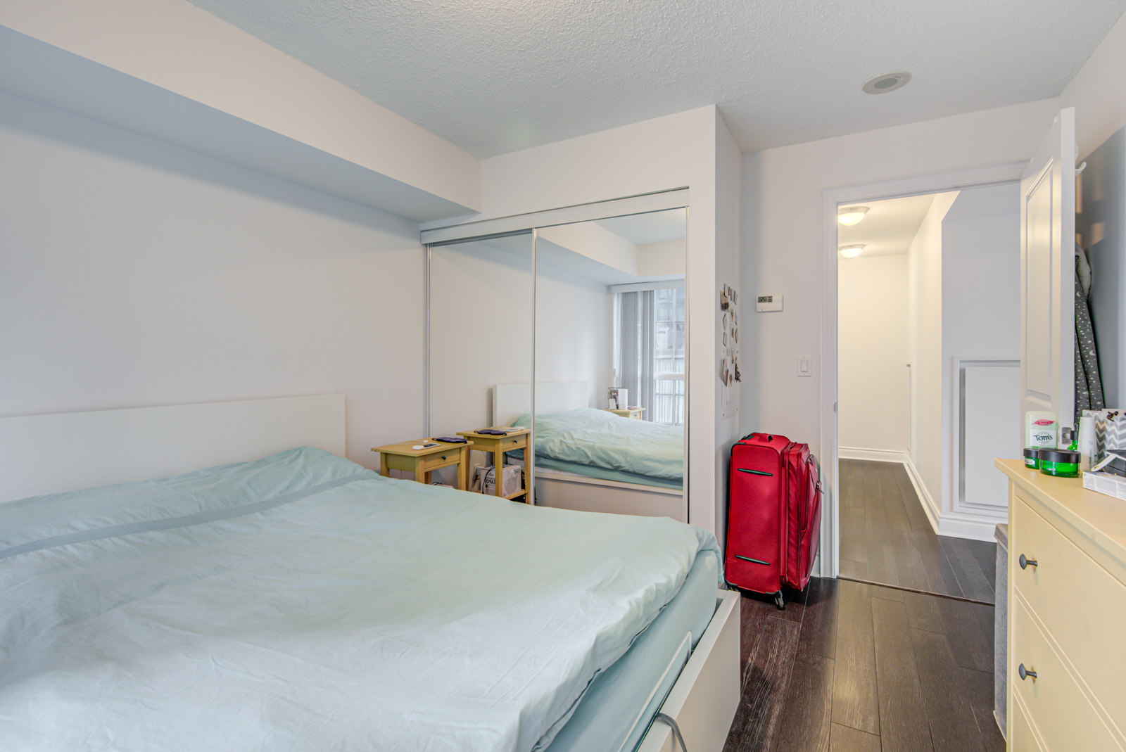 Bedroom with large white bed, sleek dark brown floors, red luggage bag, and closet with mirror doors.